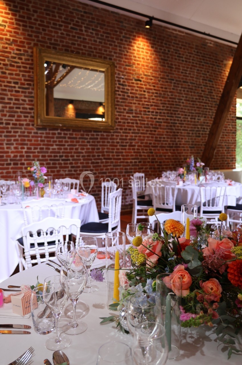 Salle de réception décorée avec des tables rondes dressées, des fleurs colorées et un mur de briques apparentes.