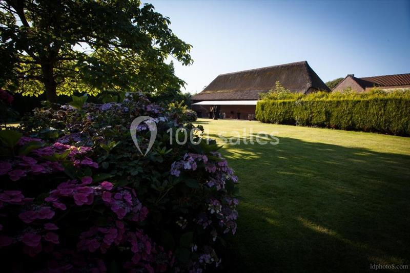 Jardin verdoyant avec des hortensias au premier plan, une pelouse bien entretenue et une grange au toit de chaume en arrière…