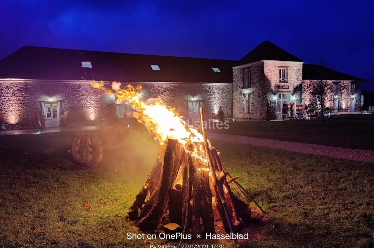 Feu de camp allumé sur une pelouse devant des bâtiments en pierre éclairés dans un environnement nocturne.