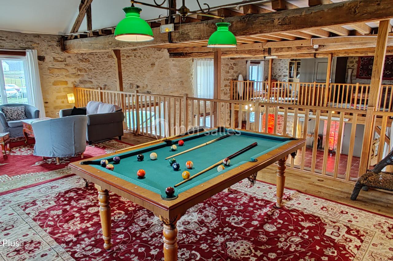 Salle avec billard, poutres apparentes, tapis rouges, fauteuils et lumière naturelle entrant par les fenêtres.