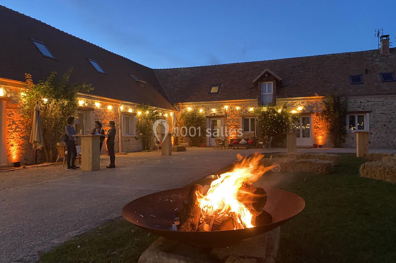 Brasero allumé dans une cour pavée entourée de bâtiments en pierre éclairés par des guirlandes lumineuses.