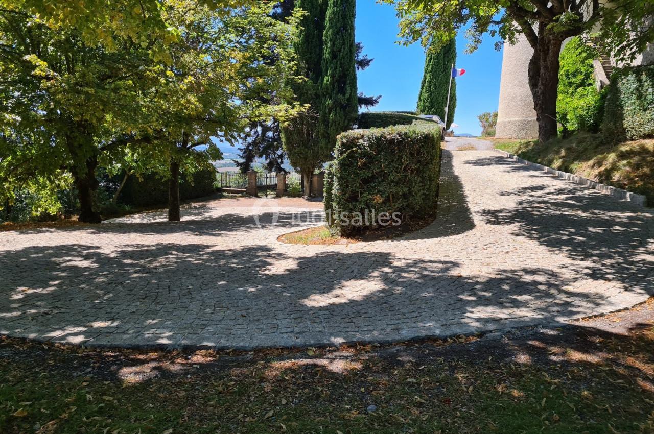 Allée pavée bordée d'arbres et de haies, menant à une tour en pierre sous un ciel bleu.