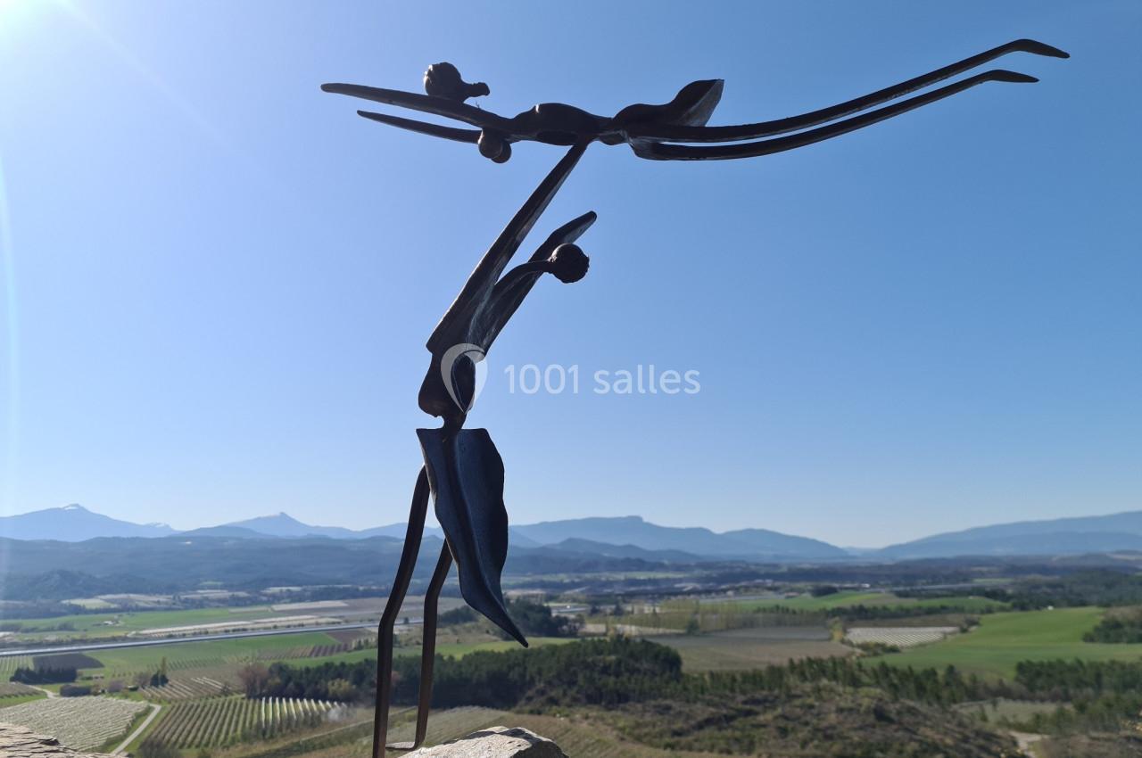 Sculpture en métal représentant deux figures élancées dansant, avec un paysage vallonné et ensoleillé en arrière-plan.