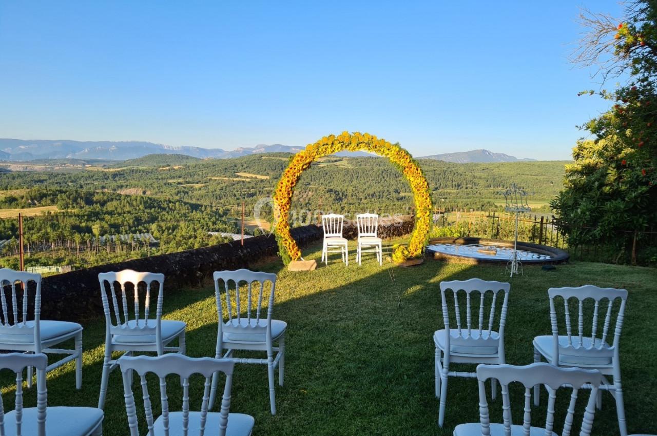 Chaises blanches disposées devant une arche fleurie sur une pelouse, avec vue sur un paysage vallonné sous un ciel dégagé.