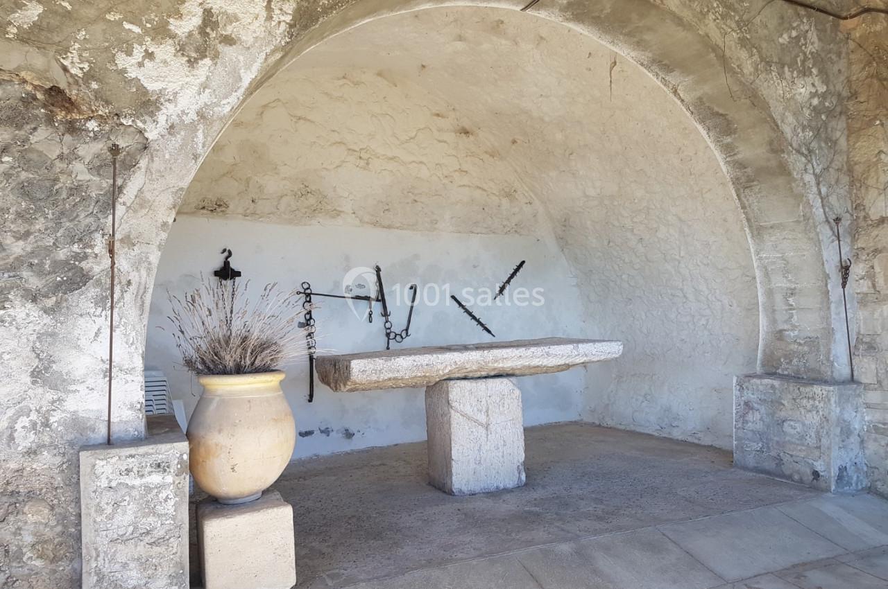 Table en pierre sous une voûte en pierre, avec un pot de fleurs et des outils anciens accrochés au mur.