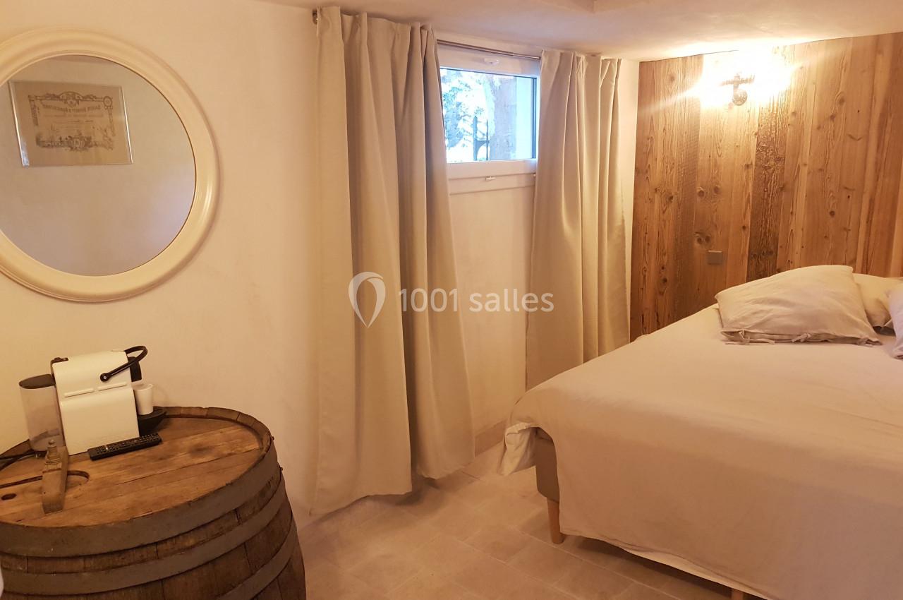 Chambre lumineuse avec lit double, murs en bois, rideaux beiges, miroir rond et table en tonneau avec machine à café.