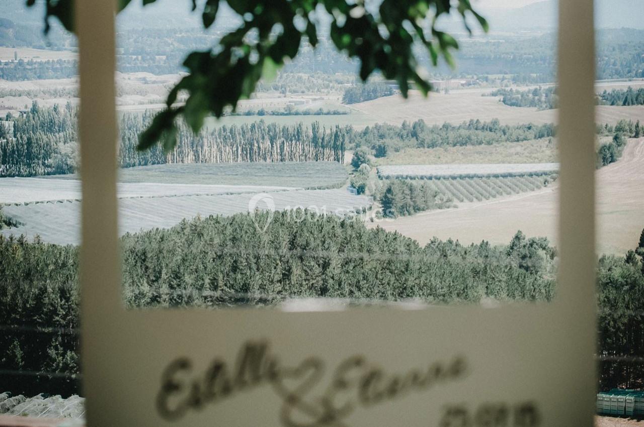 Vue d'un paysage rural avec champs, arbres et collines, encadrée par une structure blanche au premier plan.