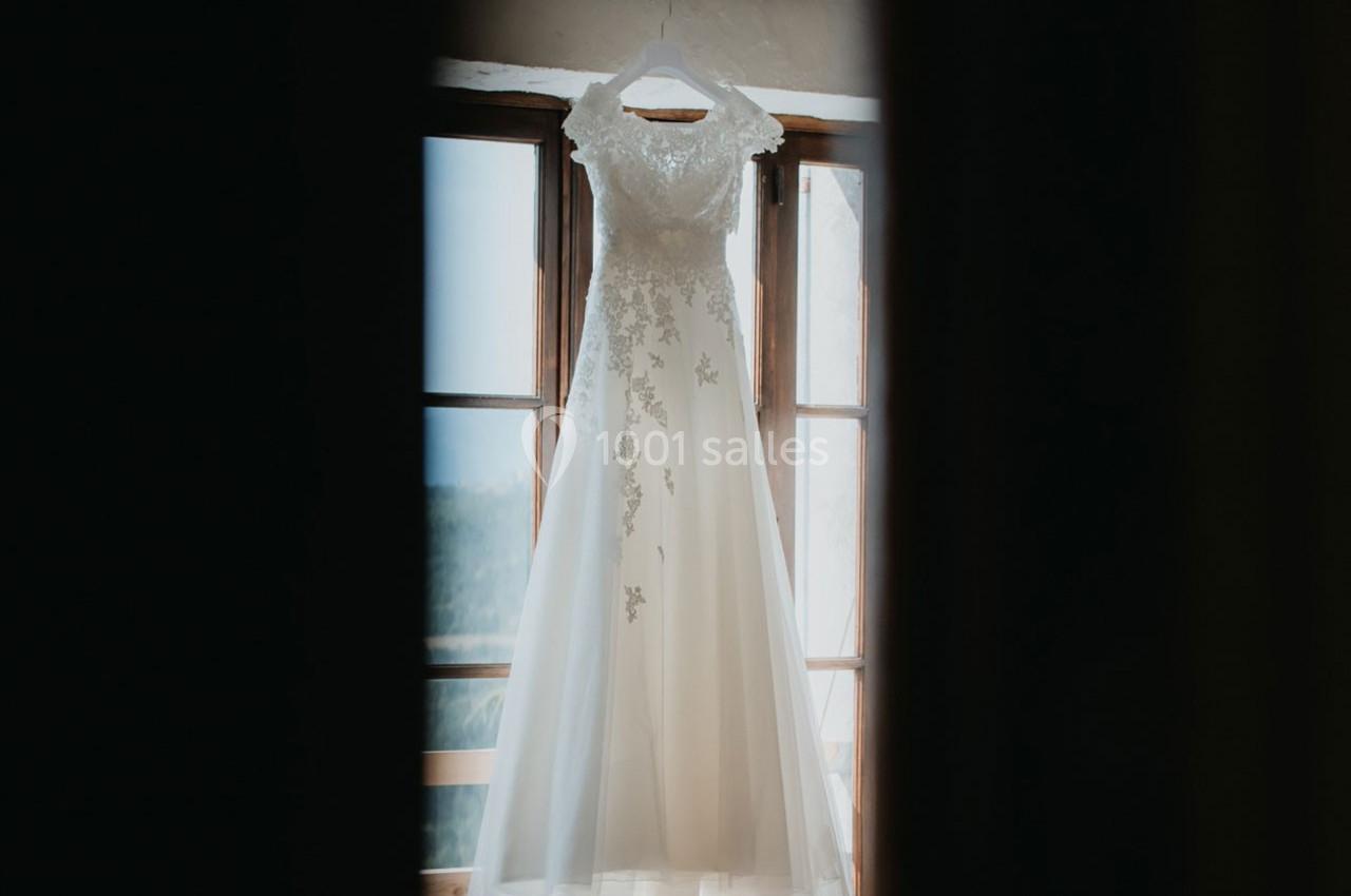 Robe de mariée blanche ornée de dentelle suspendue devant une fenêtre lumineuse.