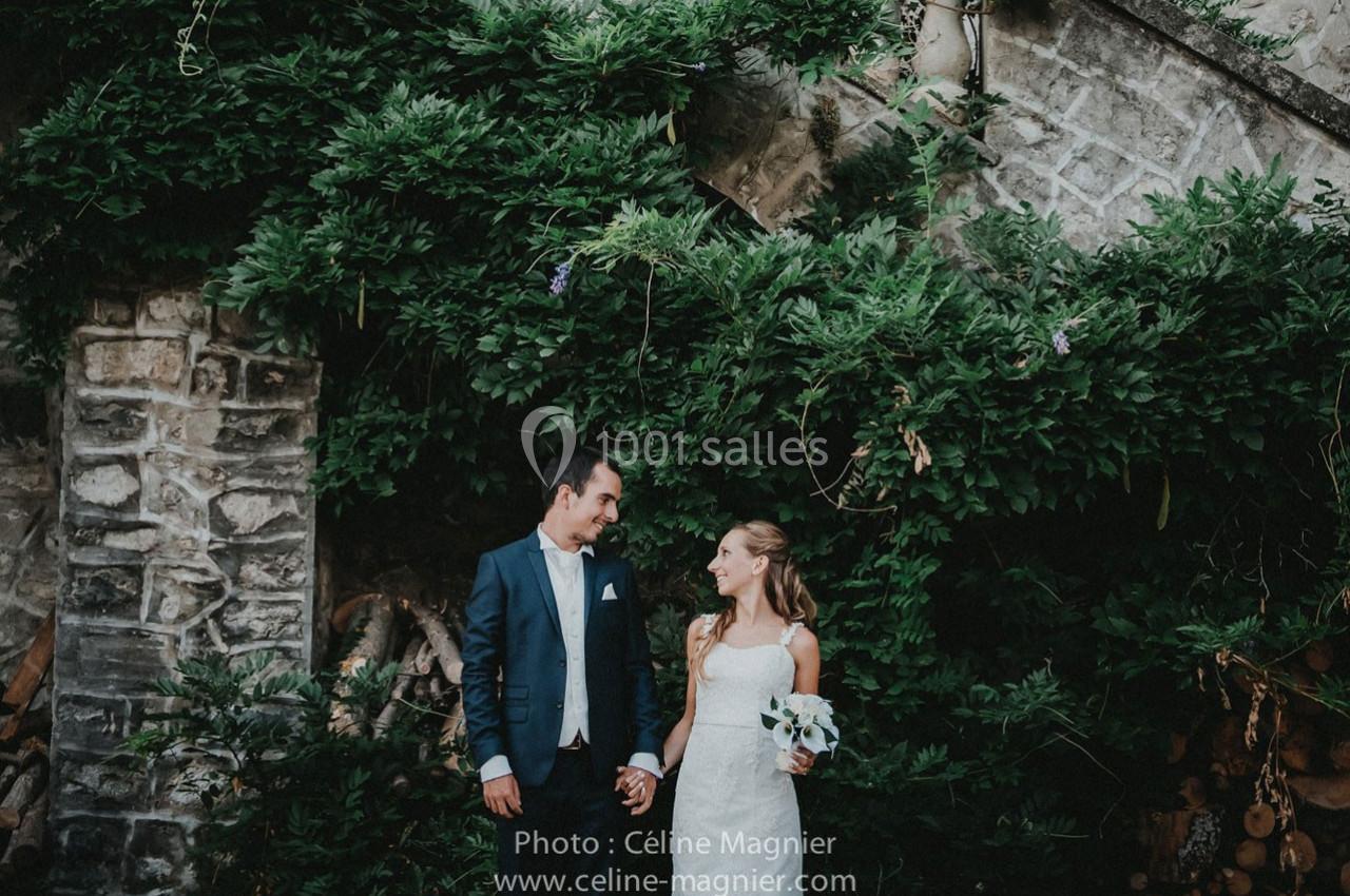 Un couple en tenue de mariage se tient la main devant un mur de pierre recouvert de verdure.