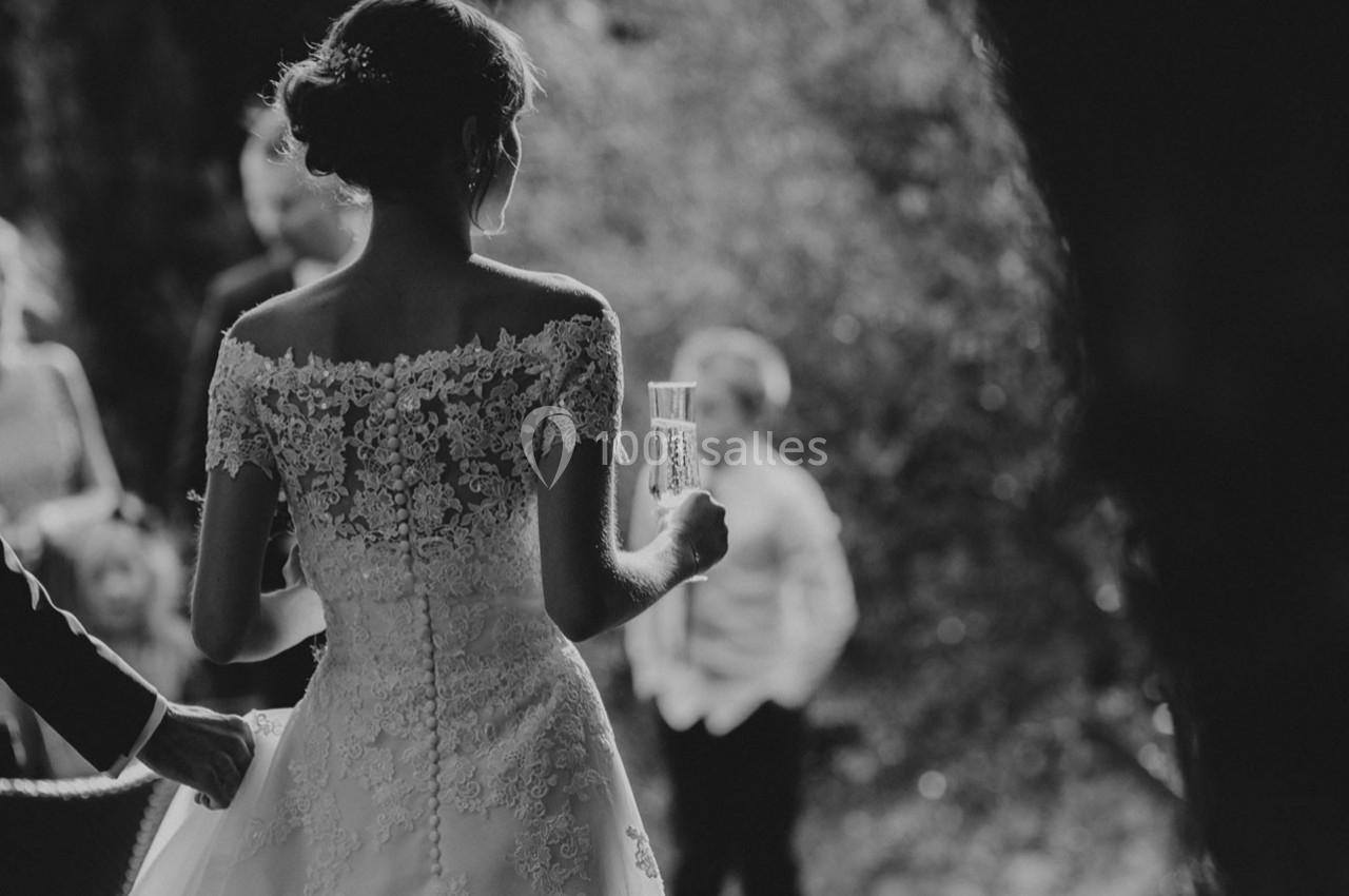 Une mariée en robe blanche ornée de dentelle tient un verre, vue de dos dans un cadre extérieur flou.