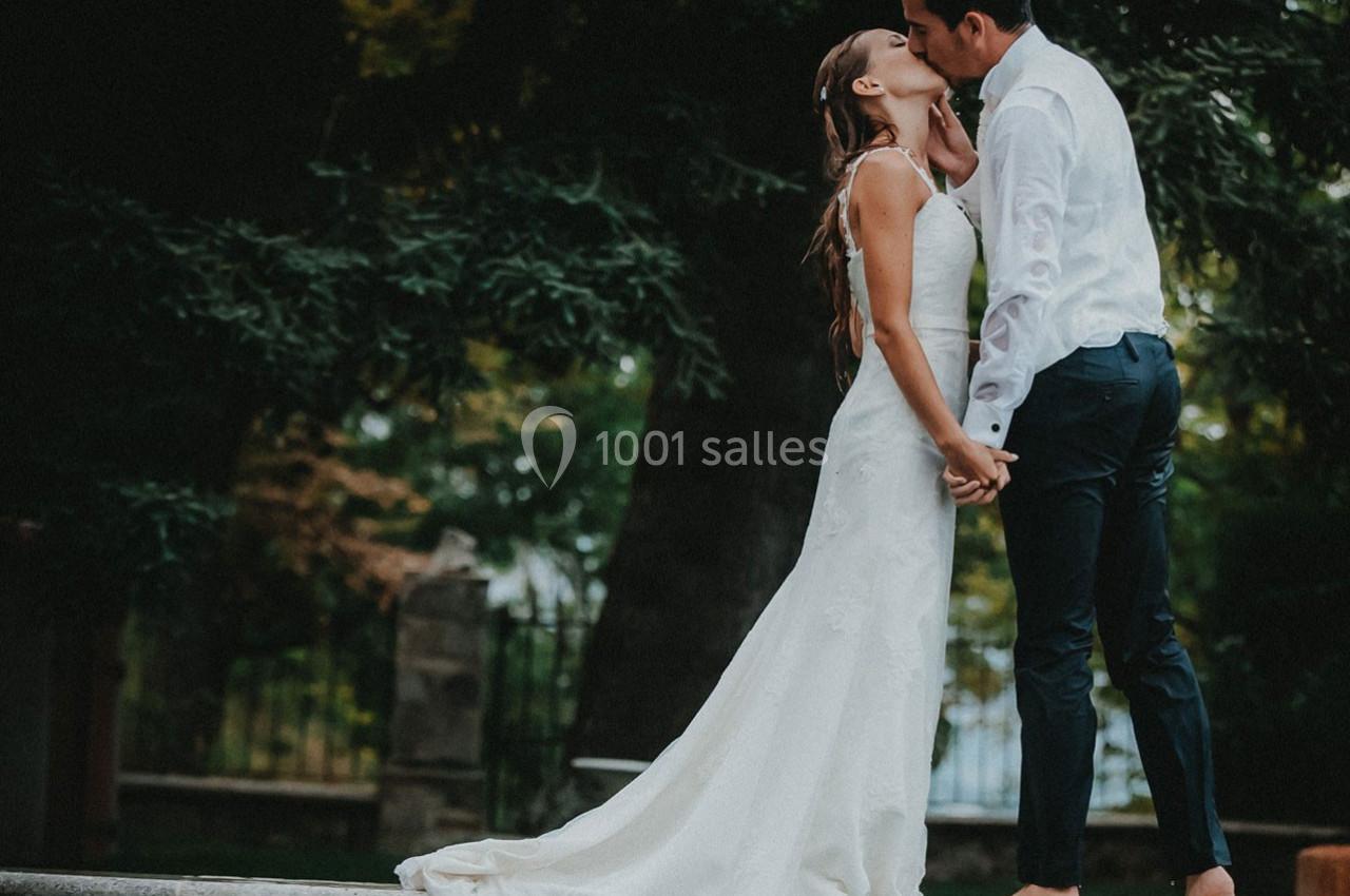 Un couple en tenue de mariage s'embrasse tendrement, debout près d'un bassin, entouré de verdure.