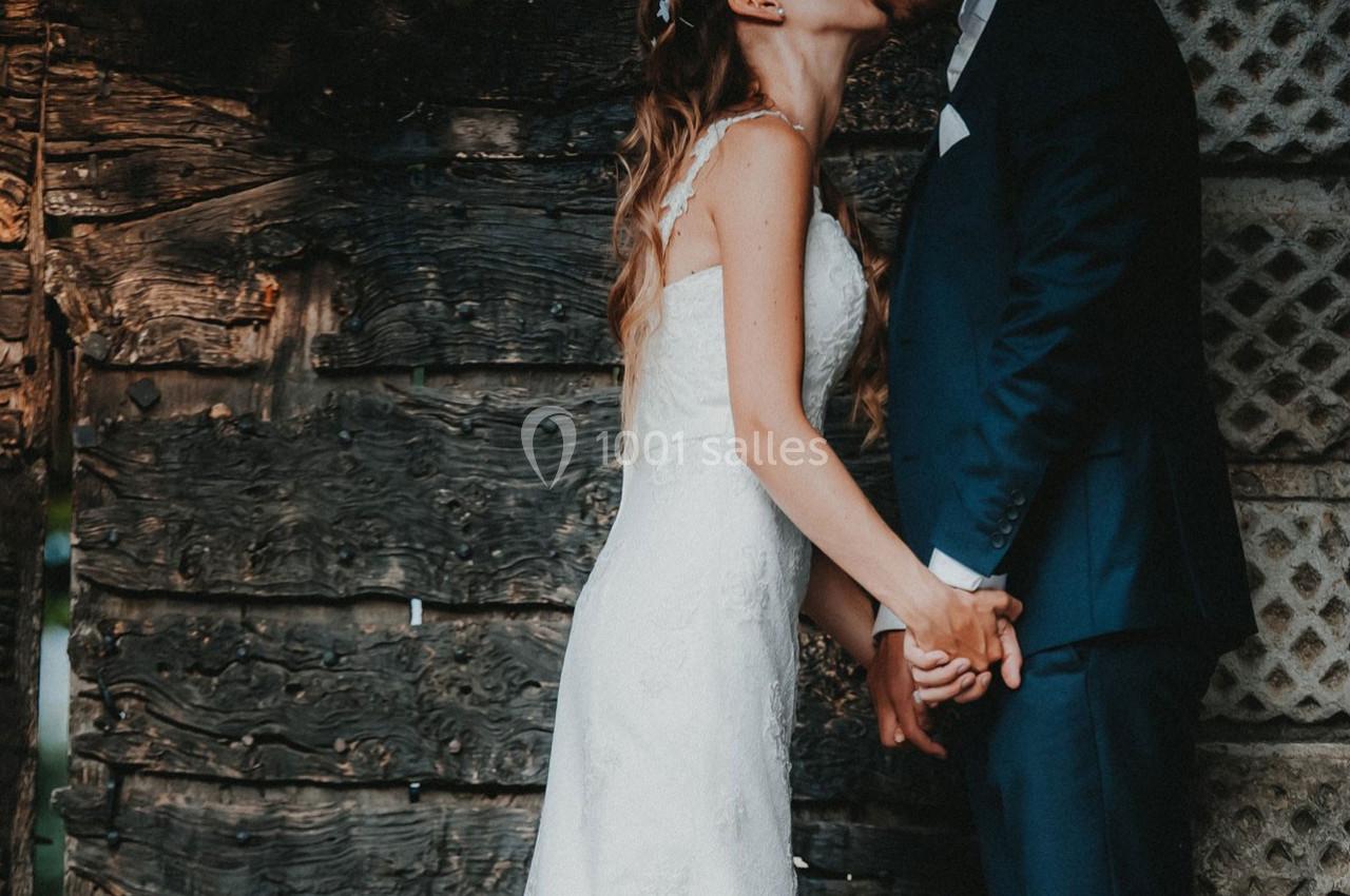 Un couple en tenue de mariage s'embrasse devant un mur en bois rustique, se tenant les mains.