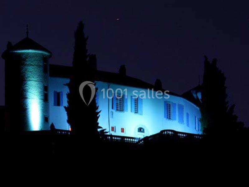 Bâtiment éclairé en bleu avec une tour ronde, entouré d'arbres, visible de nuit.
