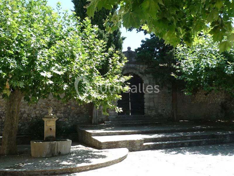 Entrée en pierre entourée d'arbres et d'ombre, avec une fontaine au premier plan dans un espace pavé.