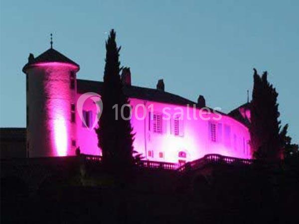 Façade d'un château éclairée en rose, entourée de cyprès, sous un ciel crépusculaire.