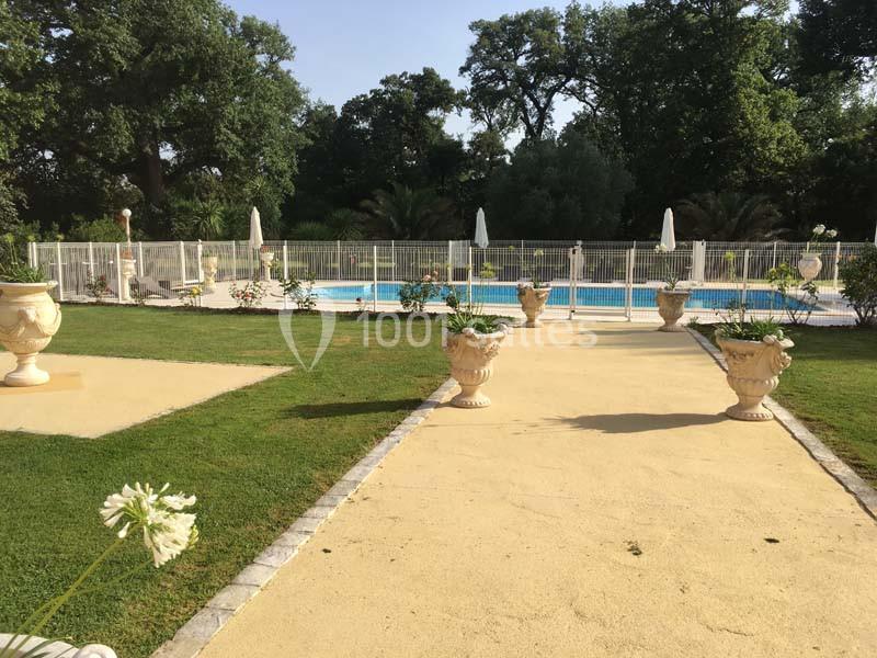 Allée bordée de pelouses et de jardinières menant à une piscine clôturée entourée de chaises longues et parasols.
