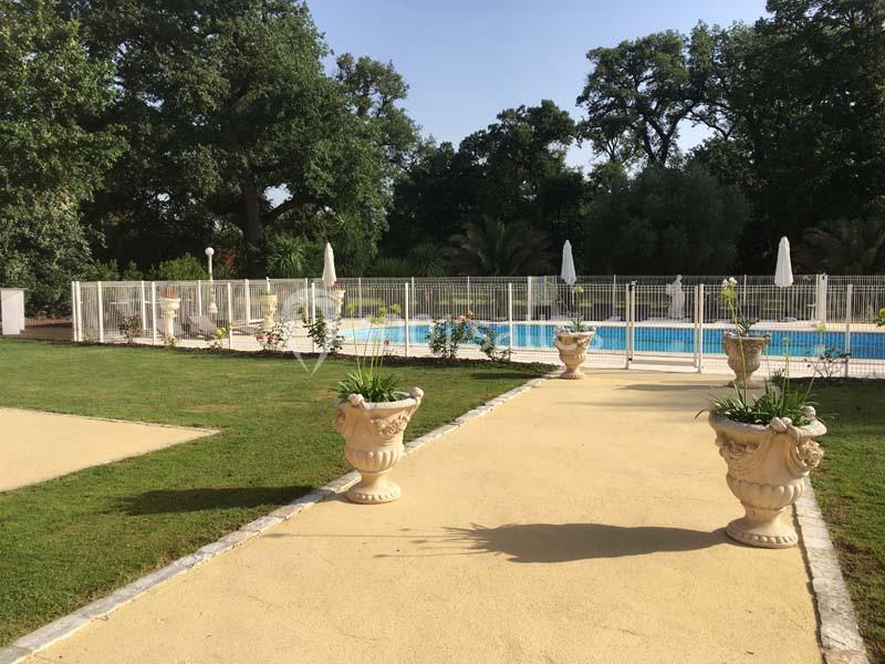 Allée bordée de pelouses menant à une piscine entourée de barrières blanches, avec des arbres en arrière-plan.