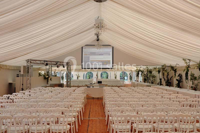 Salle de conférence sous une tente avec des rangées de chaises blanches, un écran de projection et des plantes décoratives.