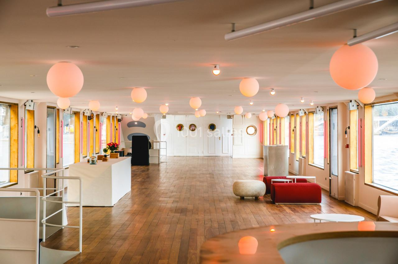 Salle lumineuse avec parquet en bois, grandes fenêtres, mobilier moderne et suspensions rondes au plafond.