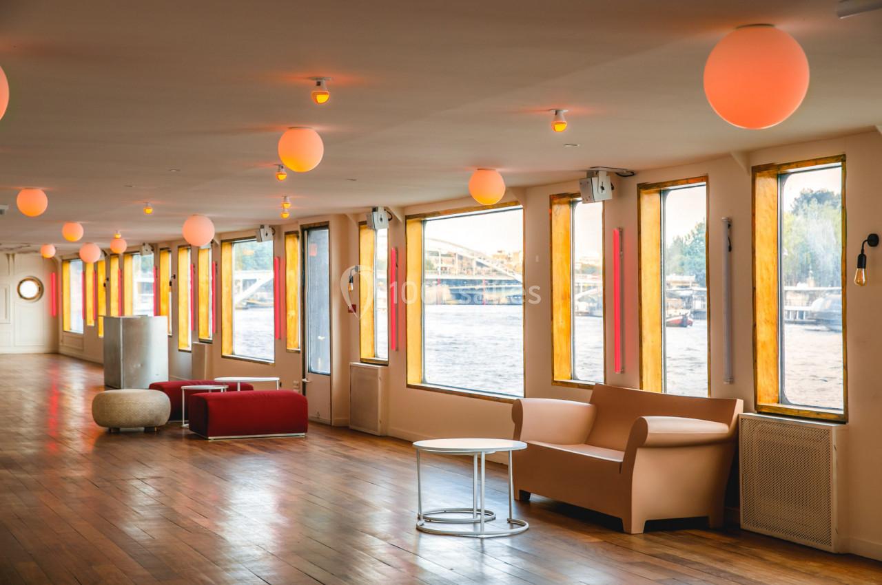 Intérieur lumineux d'une salle avec parquet, canapés, tables basses et grandes fenêtres donnant sur une rivière.