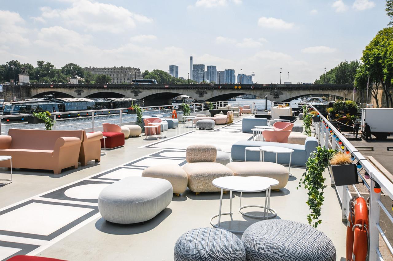 Terrasse aménagée avec des sièges et tables modernes, située au bord d'une rivière avec un pont en arrière-plan.