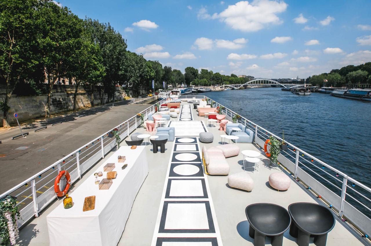 Terrasse aménagée sur une péniche avec mobilier moderne, vue sur la Seine et un pont en arrière-plan.
