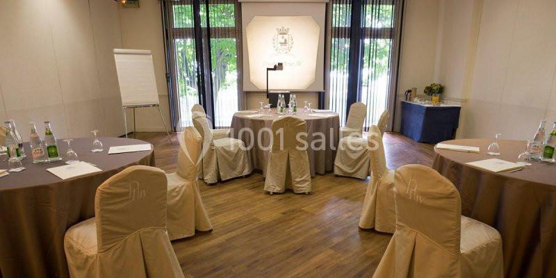 Salle de réunion lumineuse avec tables rondes, chaises couvertes, paperboard et projecteur devant une baie vitrée.