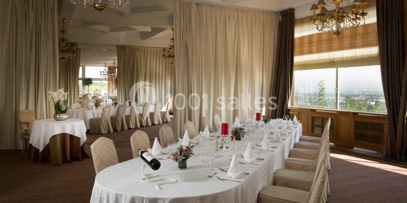 Salle de réception élégante avec tables dressées, nappes blanches, chandeliers et vue sur l'extérieur par une grande fenêtre.