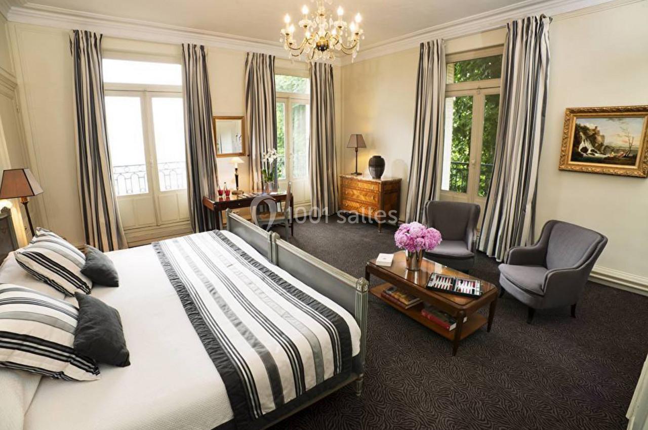 Chambre lumineuse avec lit double, fauteuils, table basse, commode, rideaux rayés et lustre au plafond.