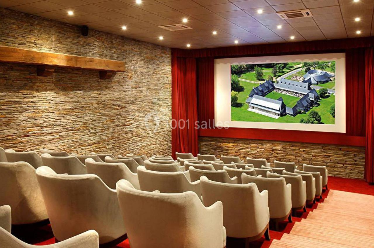 Salle de cinéma vide avec des fauteuils en velours beige, écran affichant une vue aérienne d'un bâtiment.