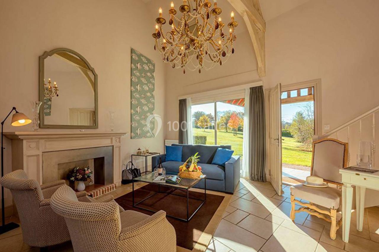Salon lumineux avec canapé bleu, fauteuils, cheminée, lustre doré et baie vitrée donnant sur un jardin verdoyant.