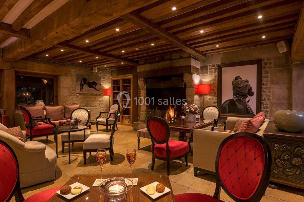 Salon chaleureux avec poutres apparentes, cheminée allumée, fauteuils rouges et éclairage tamisé.