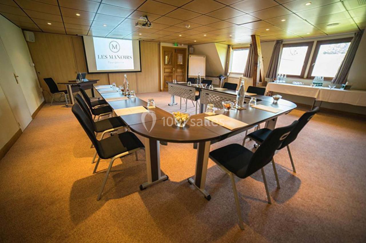 Salle de réunion lumineuse avec tables disposées en U, chaises noires, écran de projection et matériel de conférence.