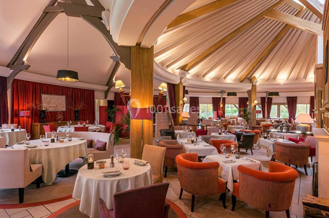 Salle de restaurant élégante avec tables rondes dressées, fauteuils colorés et plafond voûté en bois.