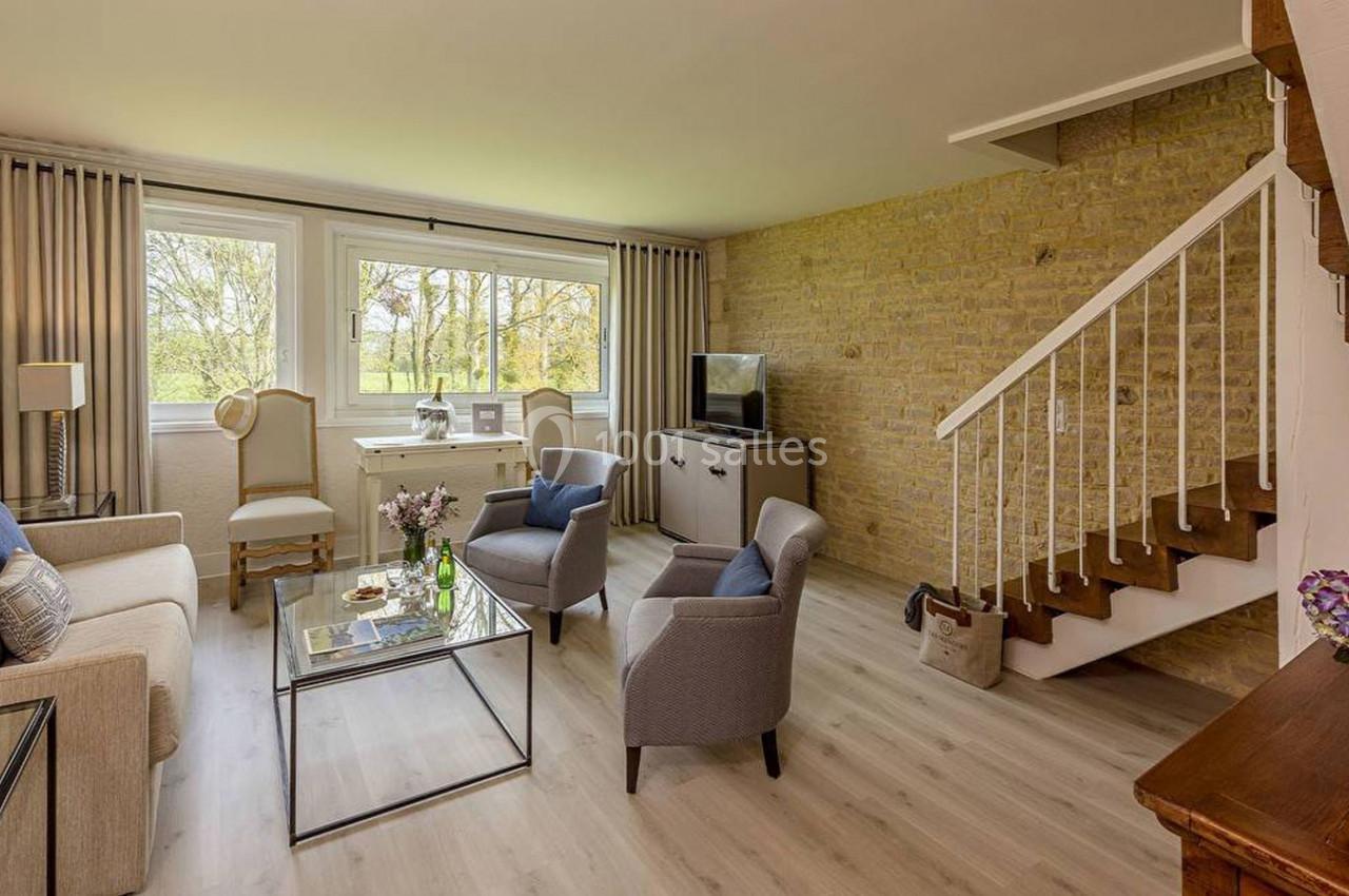 Salon lumineux avec canapé, fauteuils, table basse en verre, escalier en bois et grande fenêtre donnant sur un jardin.