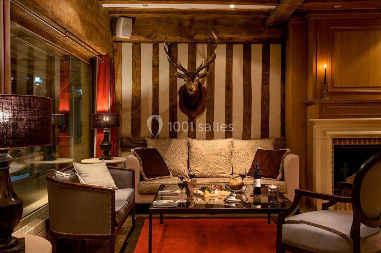 Salon chaleureux avec canapé, cheminée, tête de cerf au mur, table basse dressée et éclairage tamisé.