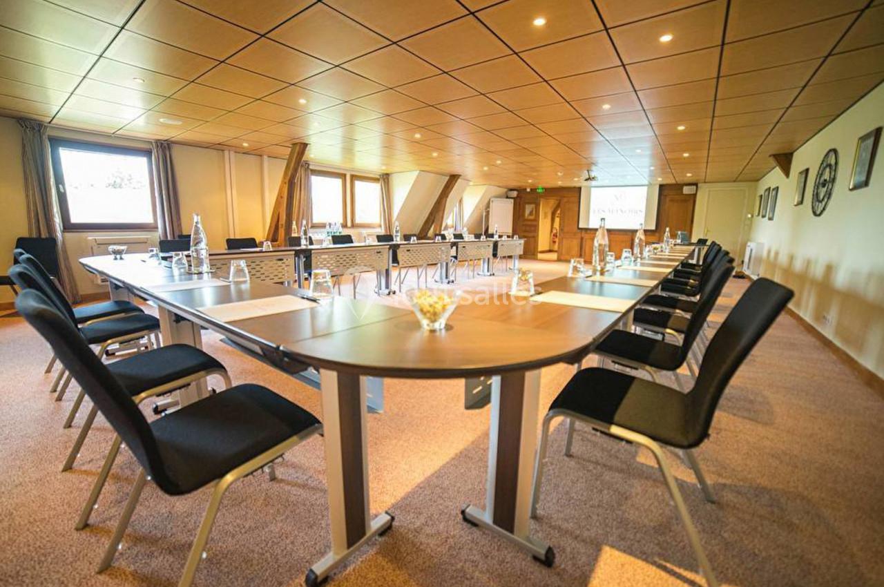 Salle de réunion lumineuse avec tables disposées en U, chaises noires et écran de projection en arrière-plan.