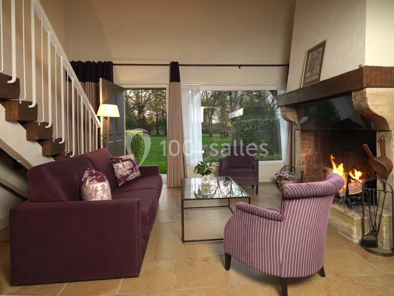 Salon chaleureux avec cheminée allumée, canapé et fauteuils violets, grande baie vitrée donnant sur un jardin.