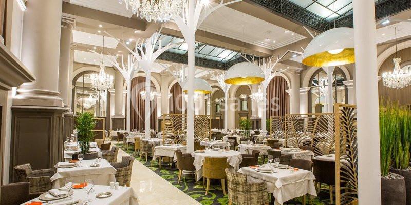 Salle de restaurant élégante avec tables dressées, chaises rembourrées, luminaires modernes et décor végétal.