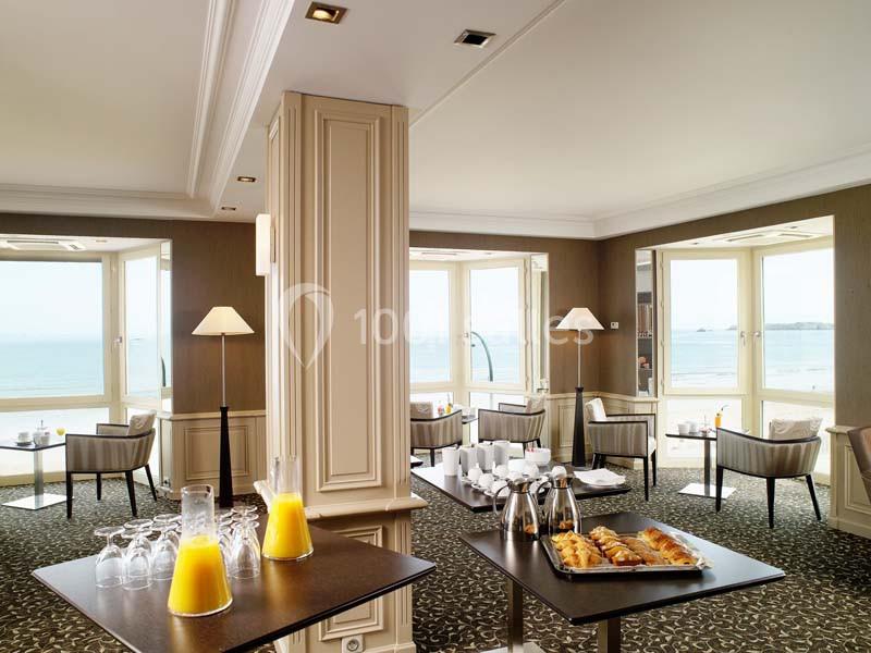 Salle lumineuse avec vue sur la mer, tables dressées pour un petit-déjeuner avec jus d'orange et viennoiseries.