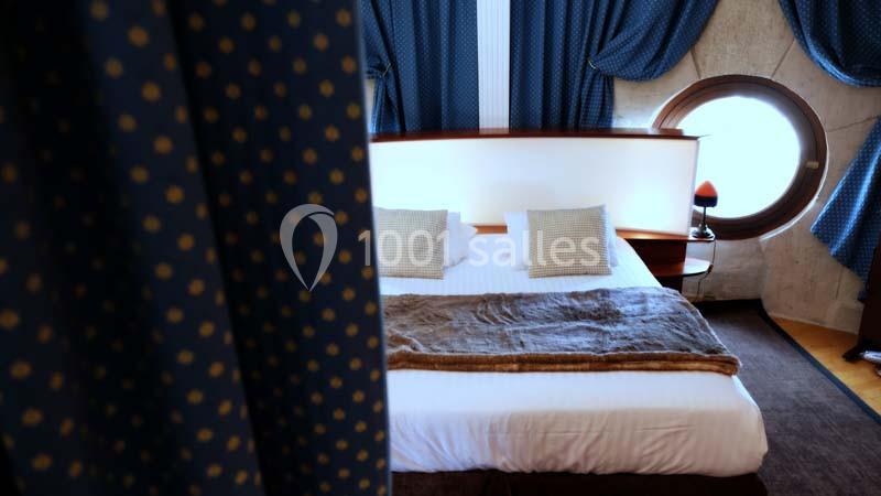 Lit double avec couverture en fourrure, deux coussins et une lampe près d'une fenêtre ronde, rideaux bleus en arrière-plan.