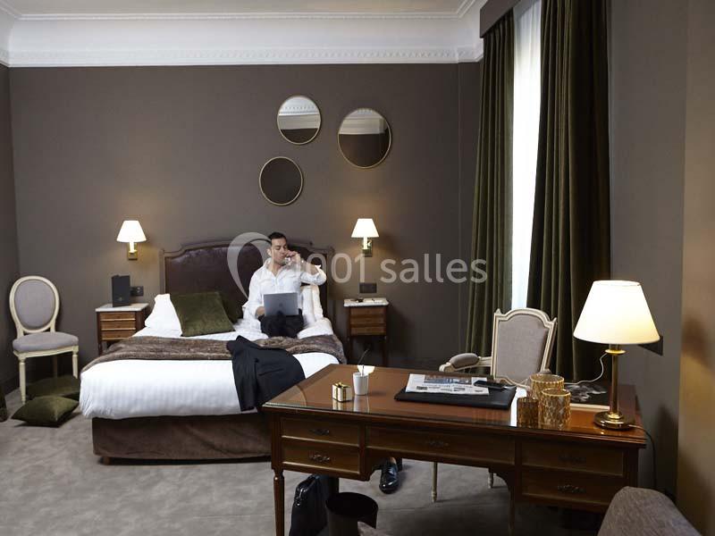 Homme assis sur un lit dans une chambre d'hôtel élégante, utilisant un ordinateur portable et parlant au téléphone.