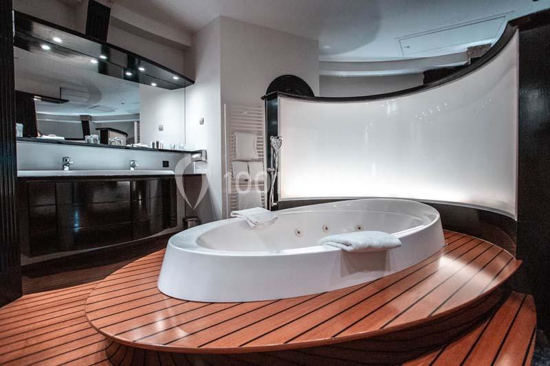 Baignoire blanche encastrée sur une plateforme en bois, avec un écran courbé et une salle de bain moderne en arrière-plan.