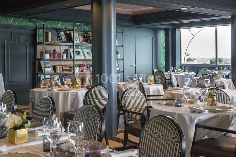 Salle de restaurant élégante avec tables dressées, chaises à motifs, étagères décoratives et grande fenêtre lumineuse.