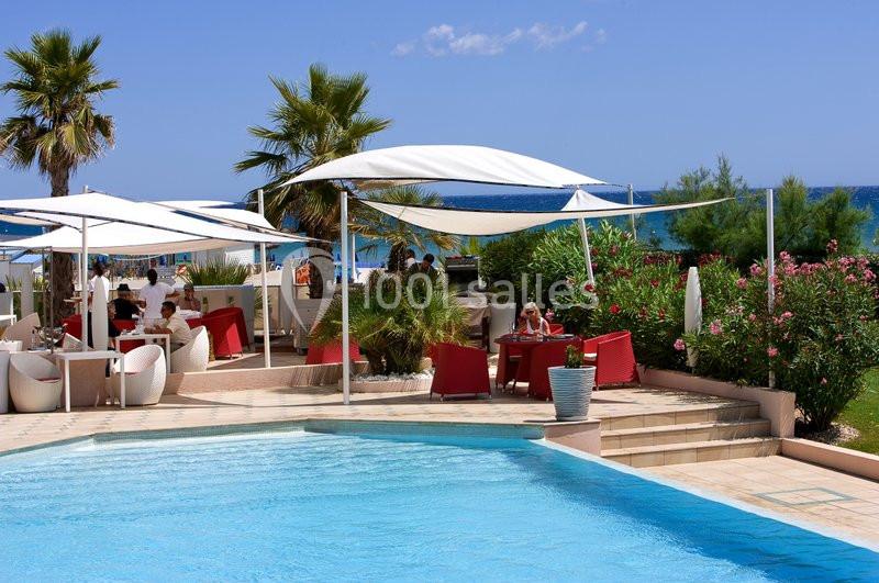 Terrasse en bord de piscine avec mobilier rouge et blanc, palmiers, parasols et vue sur la mer.