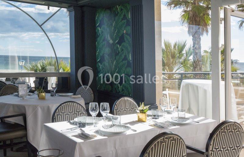 Salle de restaurant lumineuse avec vue sur la mer, tables dressées avec nappes blanches et décoration végétale.