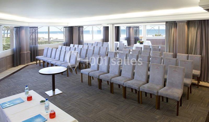 Salle de conférence lumineuse avec rangées de chaises grises, tables disposées à l'avant et vue sur l'extérieur.