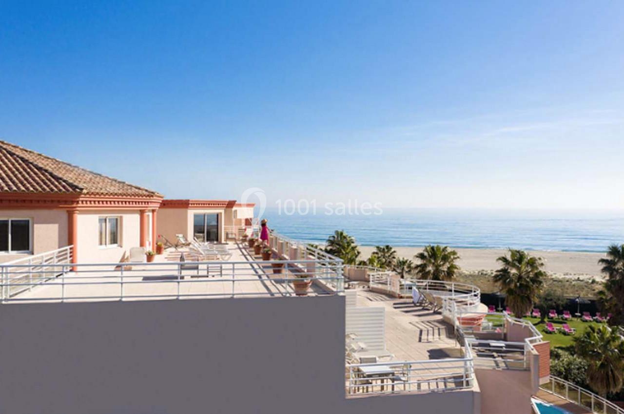 Terrasse d'un bâtiment avec vue sur la plage, des palmiers et la mer sous un ciel dégagé.