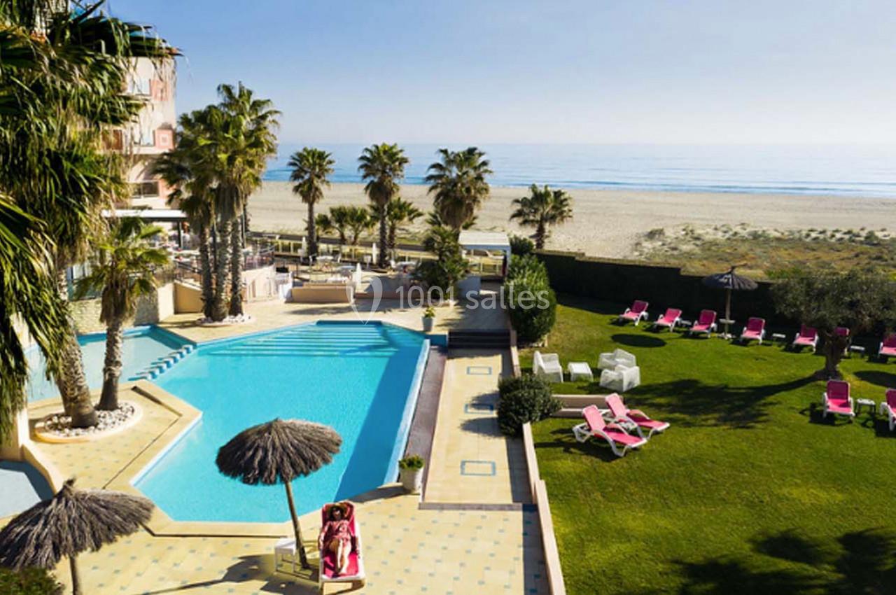 Vue d'une piscine extérieure entourée de palmiers, avec des transats et une plage en arrière-plan.