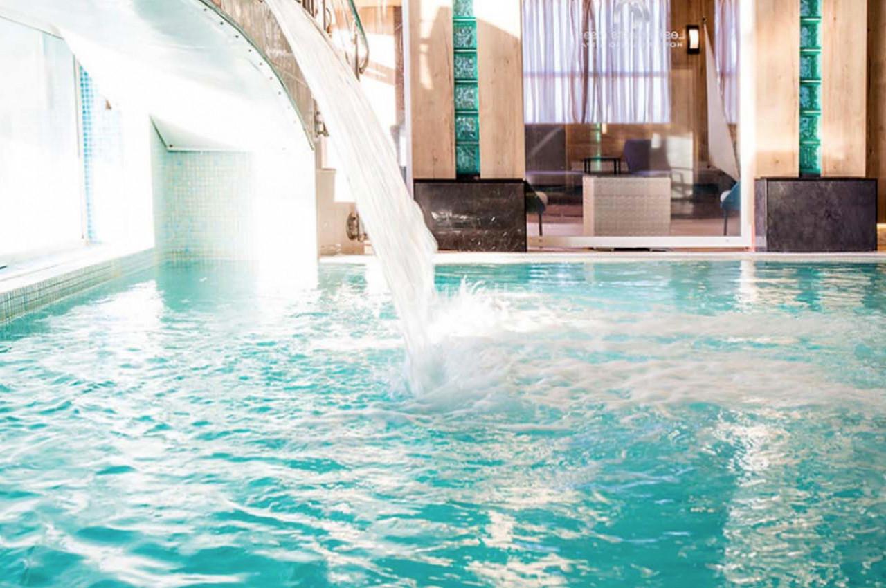 Cascade d'eau tombant dans une piscine intérieure lumineuse avec des carreaux turquoise et des baies vitrées en arrière-plan.